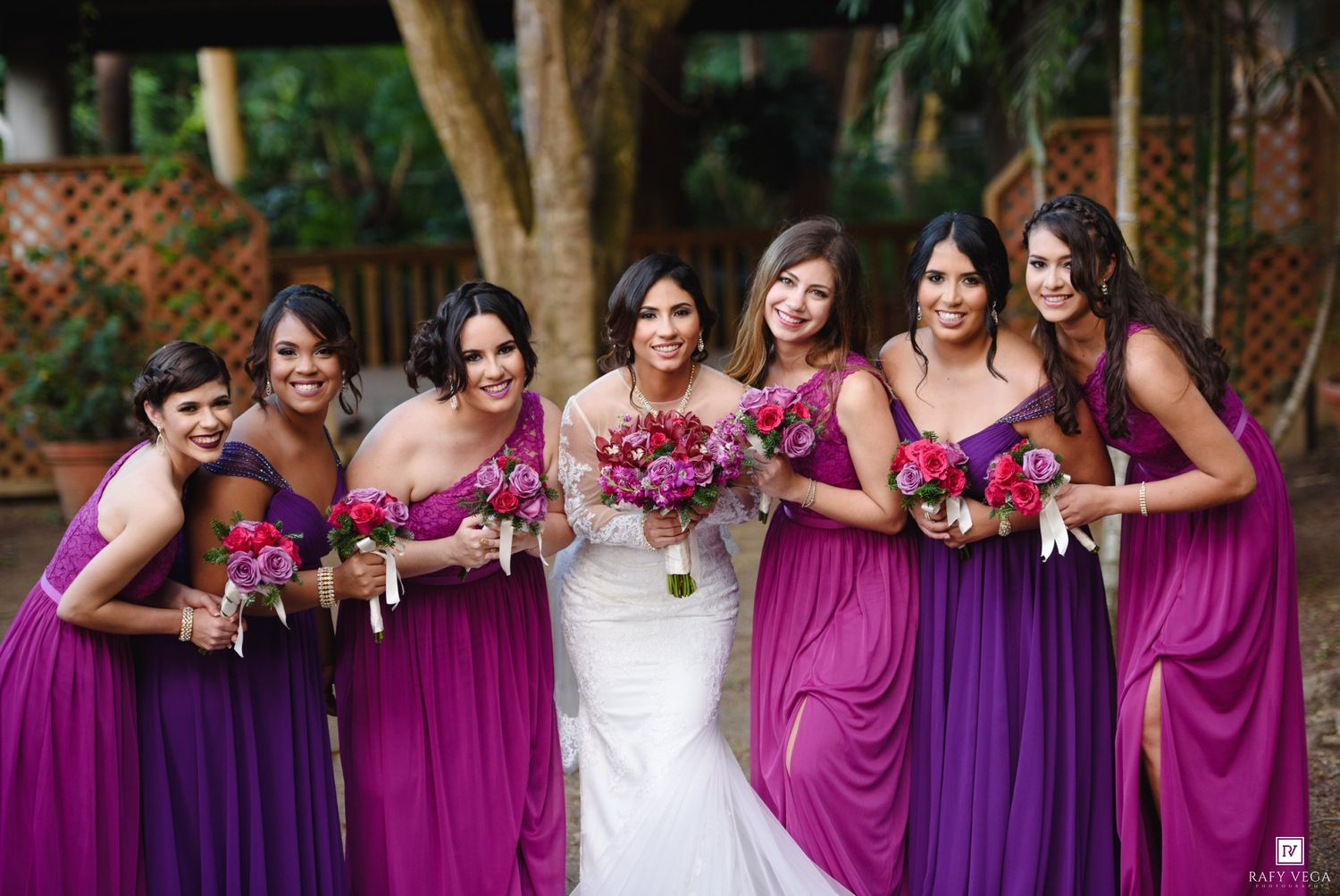 Bride in white gown poses with six bridesmaids in purple and magenta dresses holding bouquets outdoors.