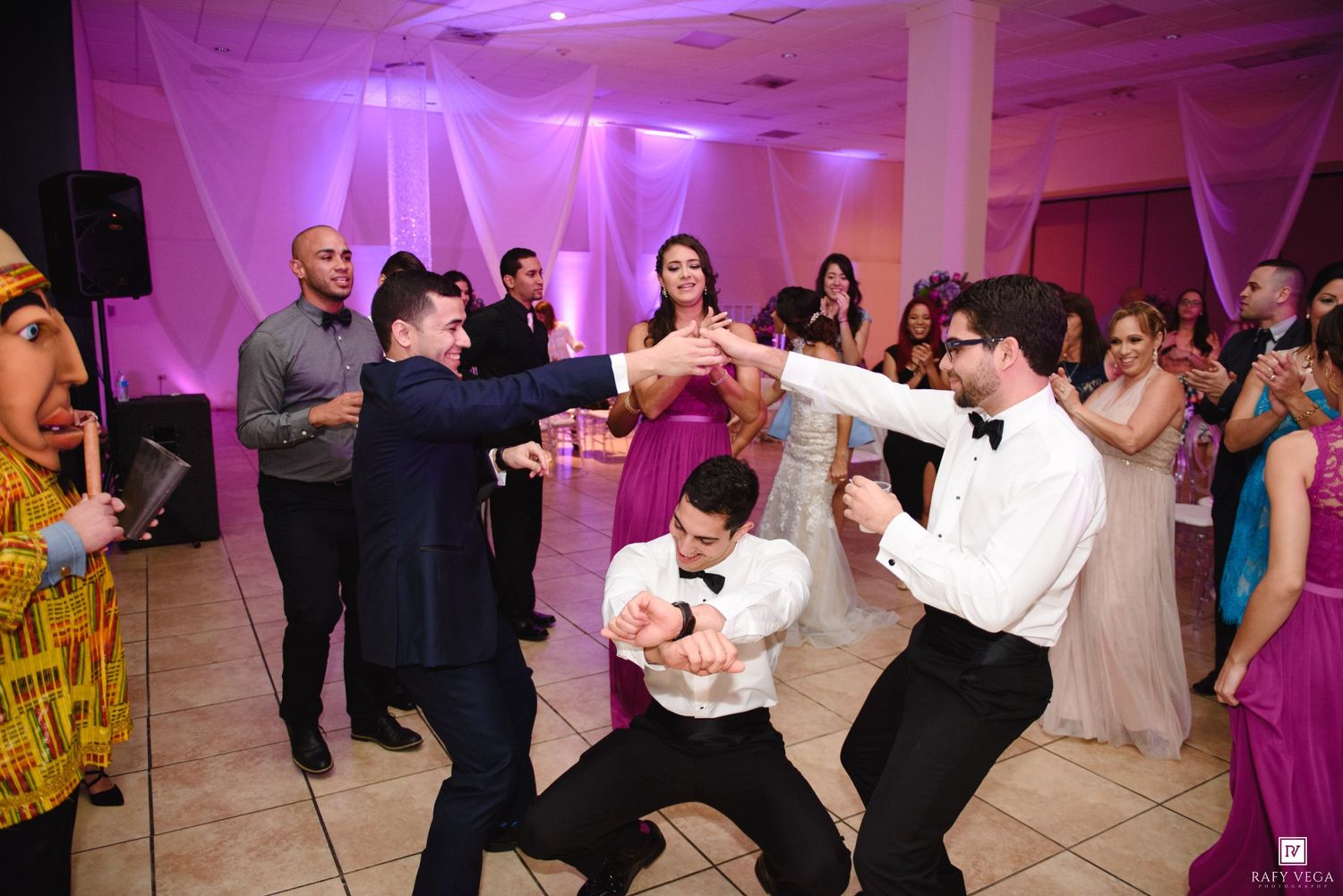Men in tuxedos dance energetically at a wedding reception with guests watching on a decorated dance floor.