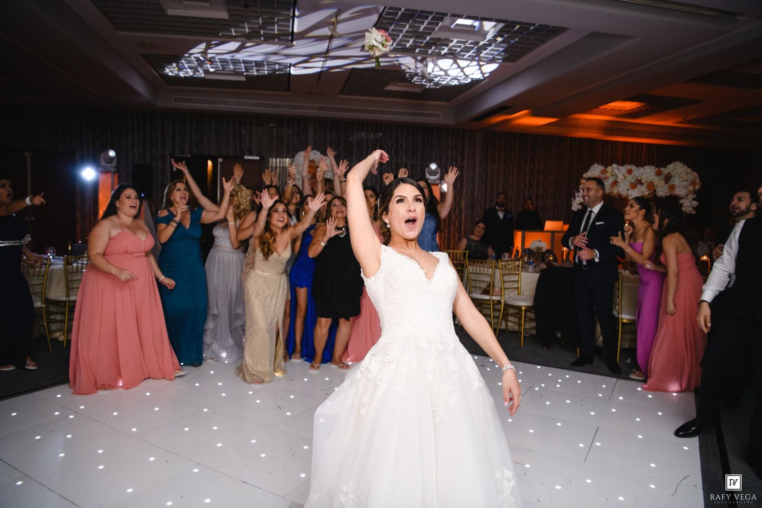 Bride tossing bouquet to excited guests on a white dance floor at wedding reception.