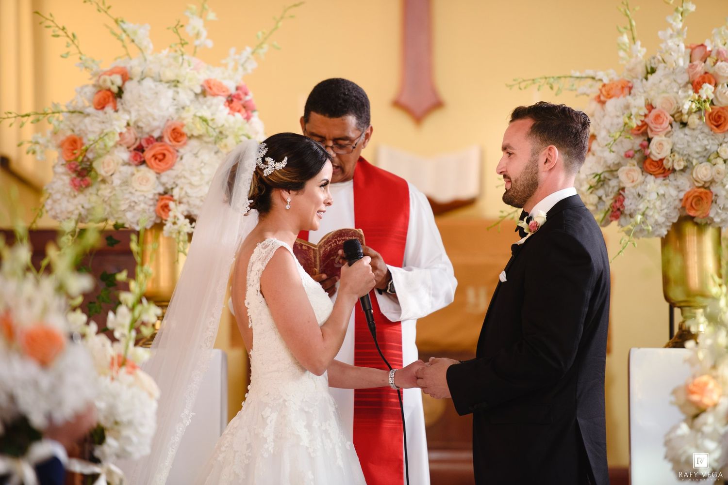 A bride and groom exchange vows at a church altar adorned with peach and white floral arrangements.