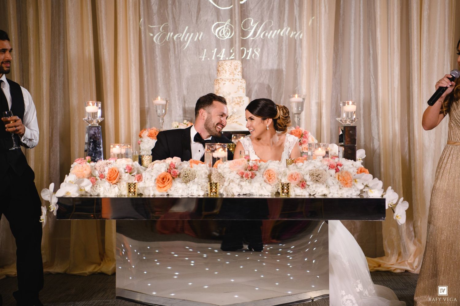 Bride and groom share a toast at elegant wedding sweetheart table adorned with peach floral arrangements and candlelight.