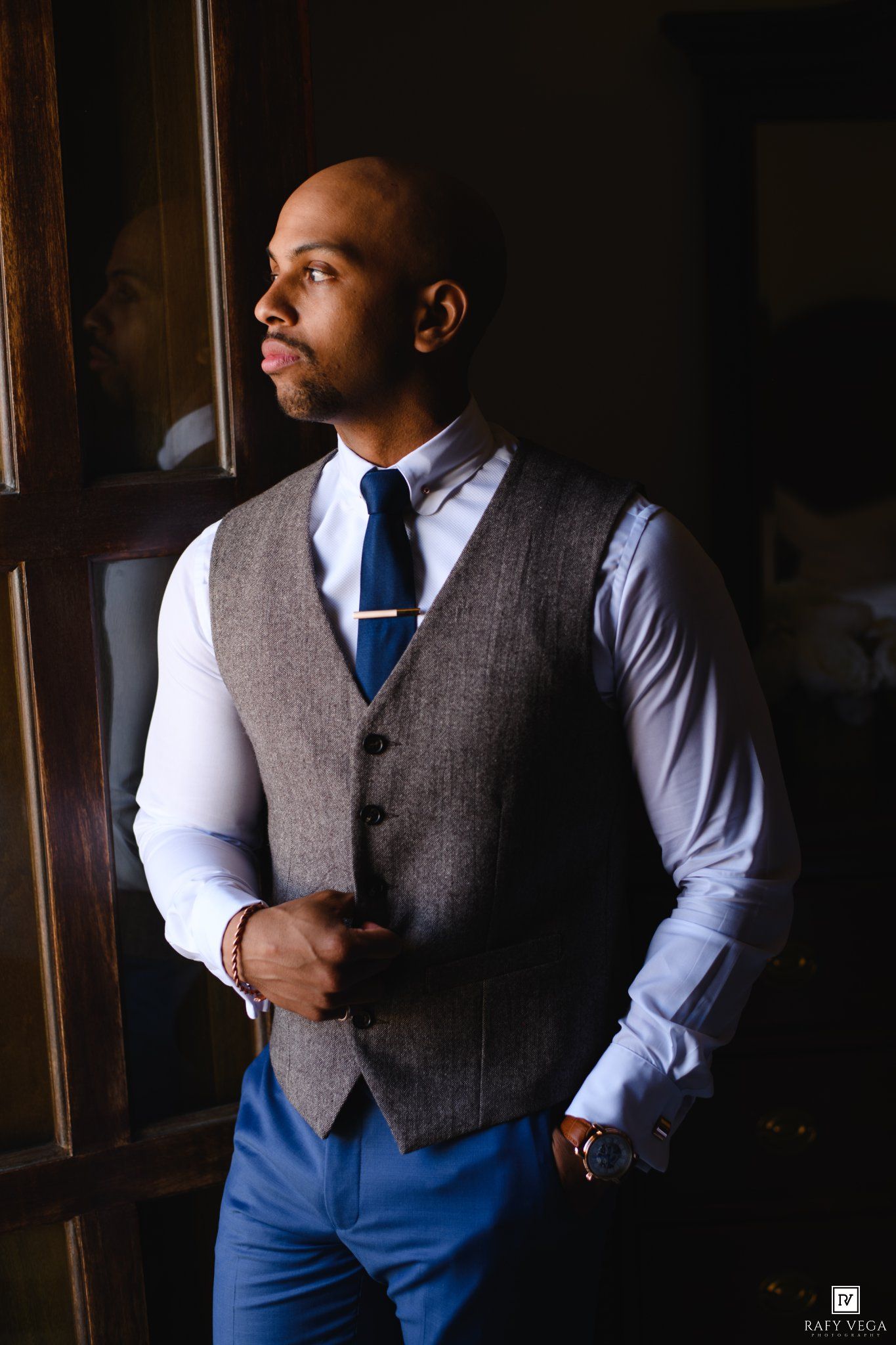 A well-dressed bald man in a gray vest, blue tie, and blue trousers gazes thoughtfully out a window.
