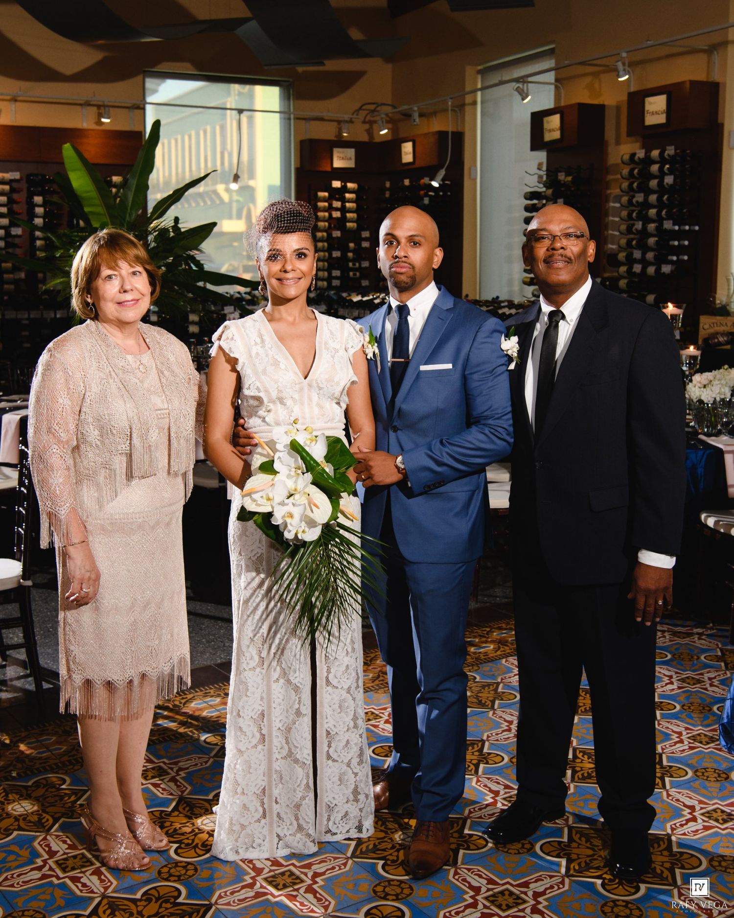 Wedding party of four posing in elegant venue, bride holding white bouquet in lace gown beside groom in blue suit.