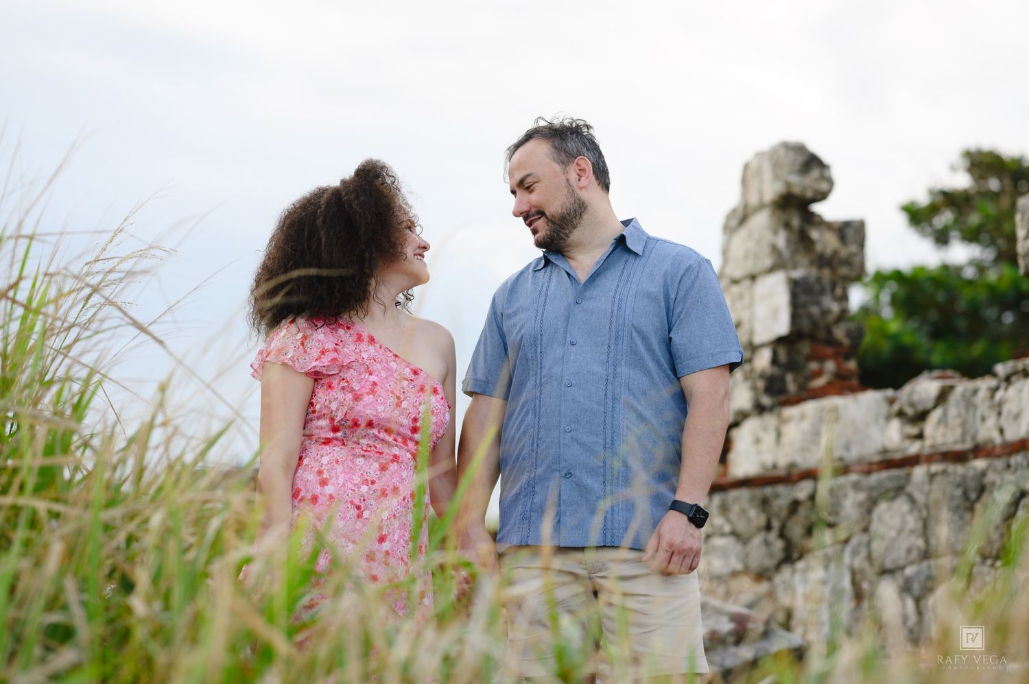Couple holding hands smiling at each other outdoors near stone ruins with tall grass in foreground.