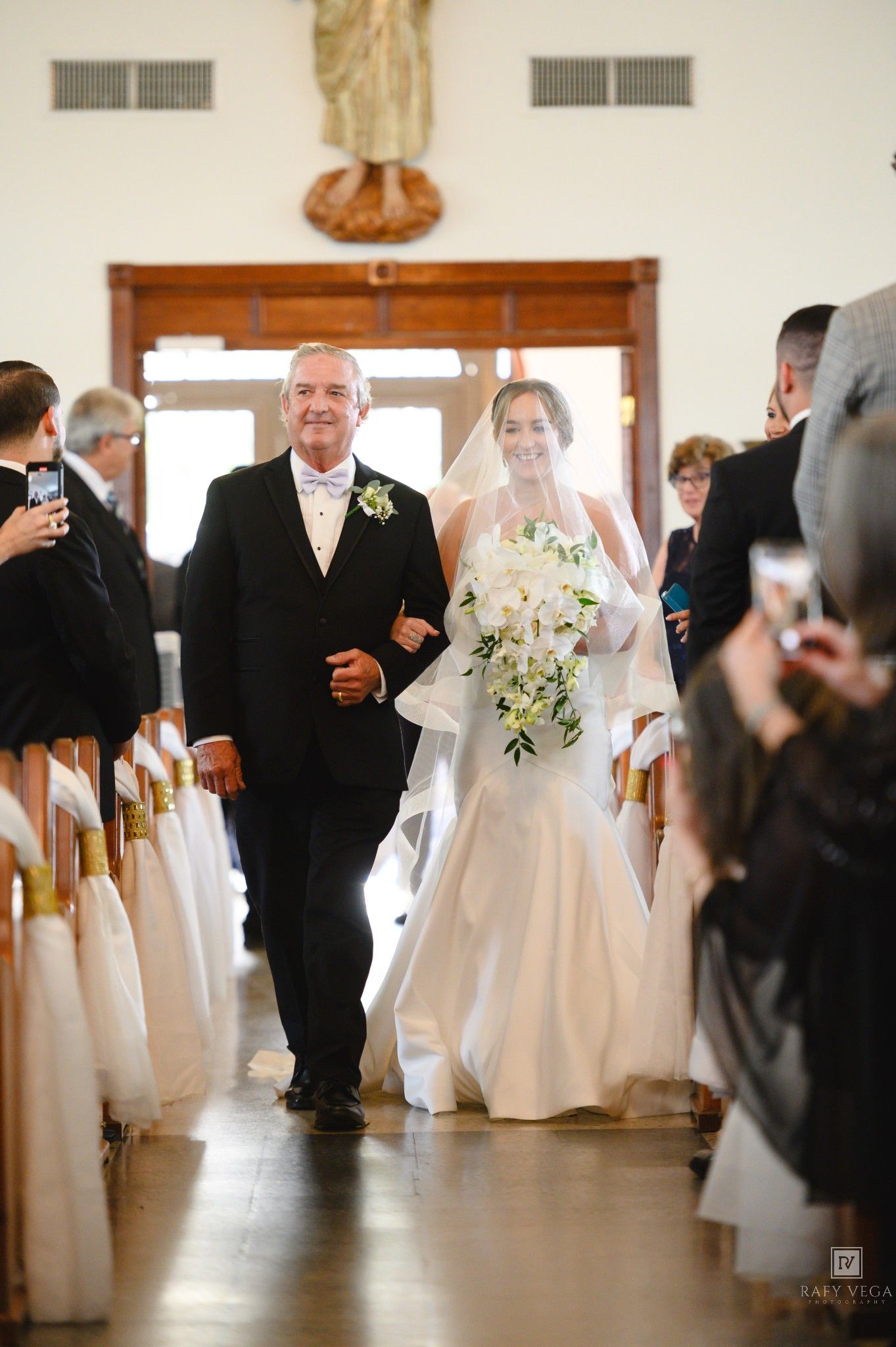 Bride and father walk down church aisle, bride holding white floral bouquet in elegant wedding gown with veil.
