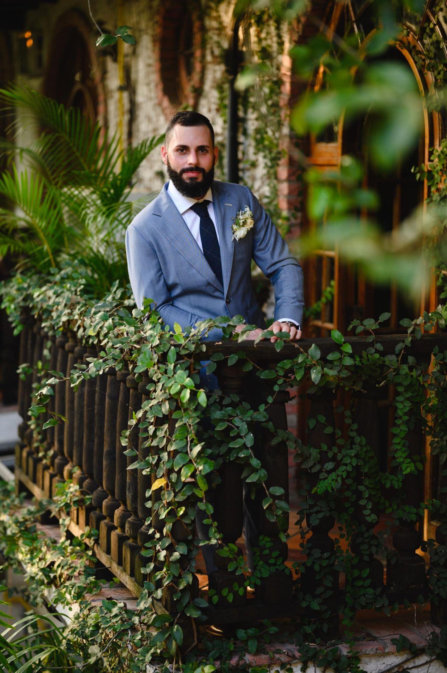 Groom in gray suit with boutonniere poses on vine-covered balcony surrounded by lush tropical greenery.