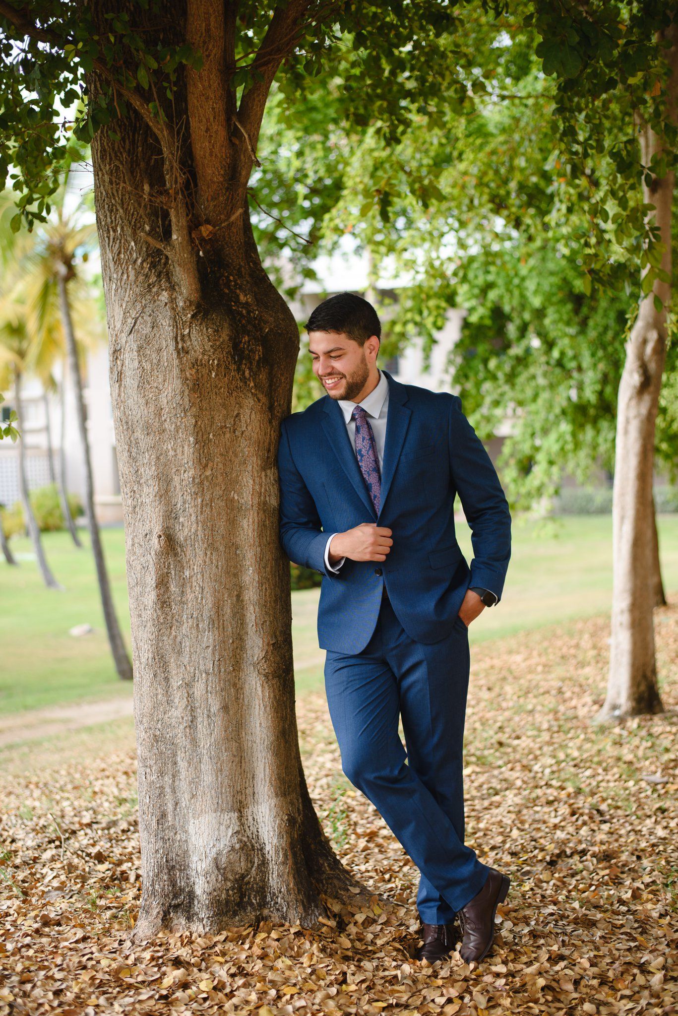 A young man in a navy blue suit and tie leans against a tree in a park, smiling downward at autumn leaves.