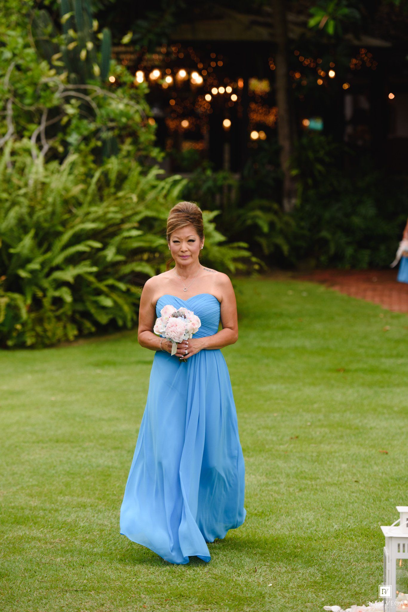 Bridesmaid in a blue strapless gown walks down an outdoor aisle holding a white bouquet at a garden wedding.