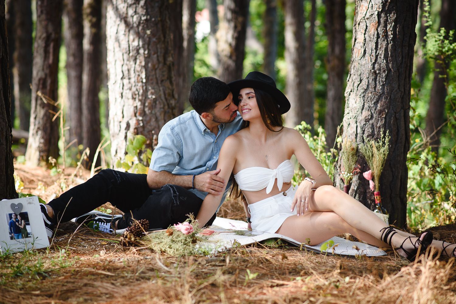 Romantic couple sharing a tender moment during a picnic in a pine forest, woman in white dress and black hat.