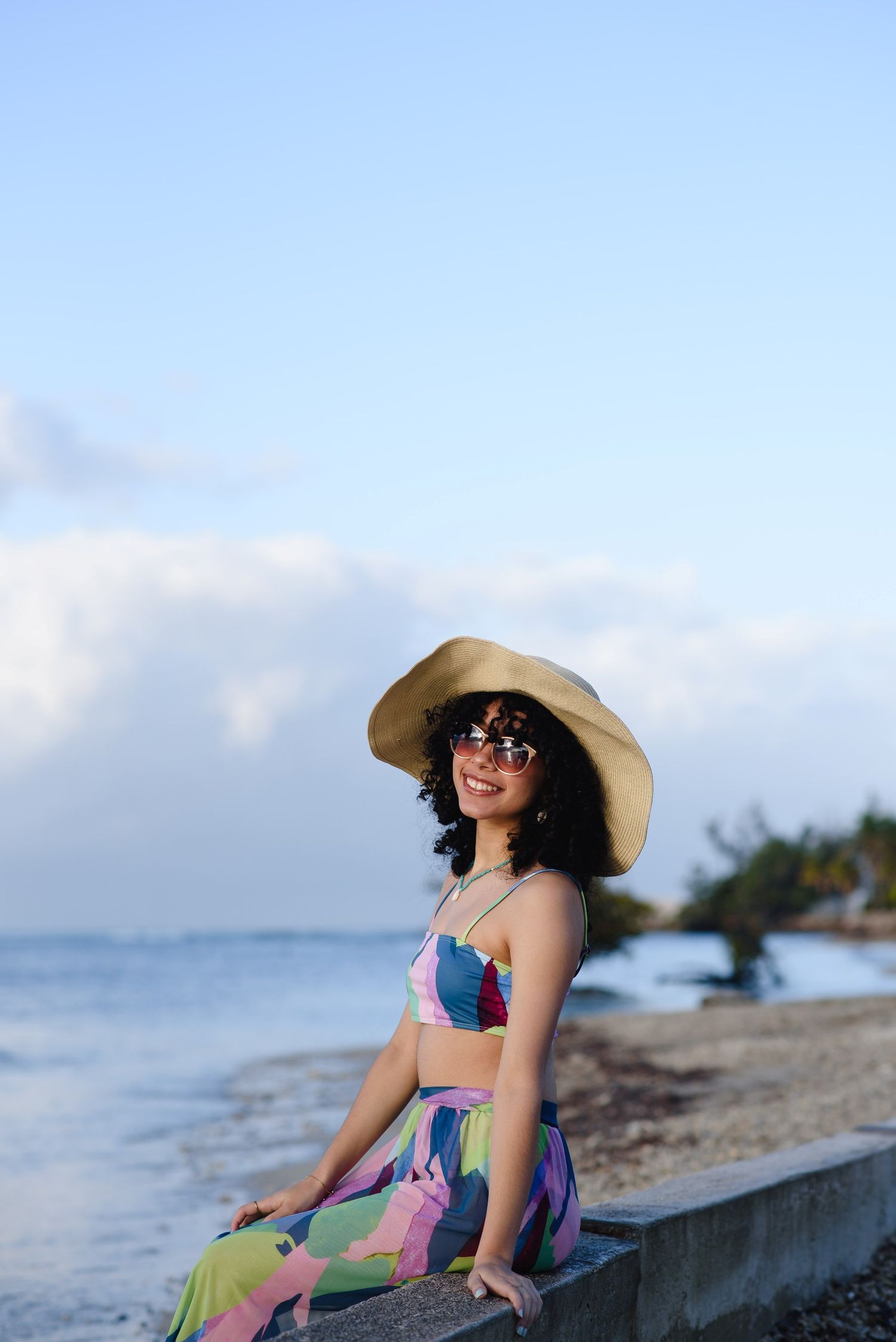 Smiling woman in colorful two-piece outfit, wide-brim hat, and sunglasses sitting on a beach wall.