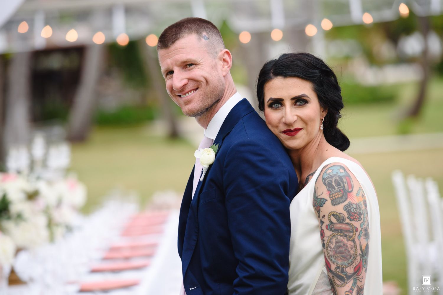 Smiling bride with tattoo sleeve and groom in navy suit pose back-to-back at elegant outdoor wedding reception.