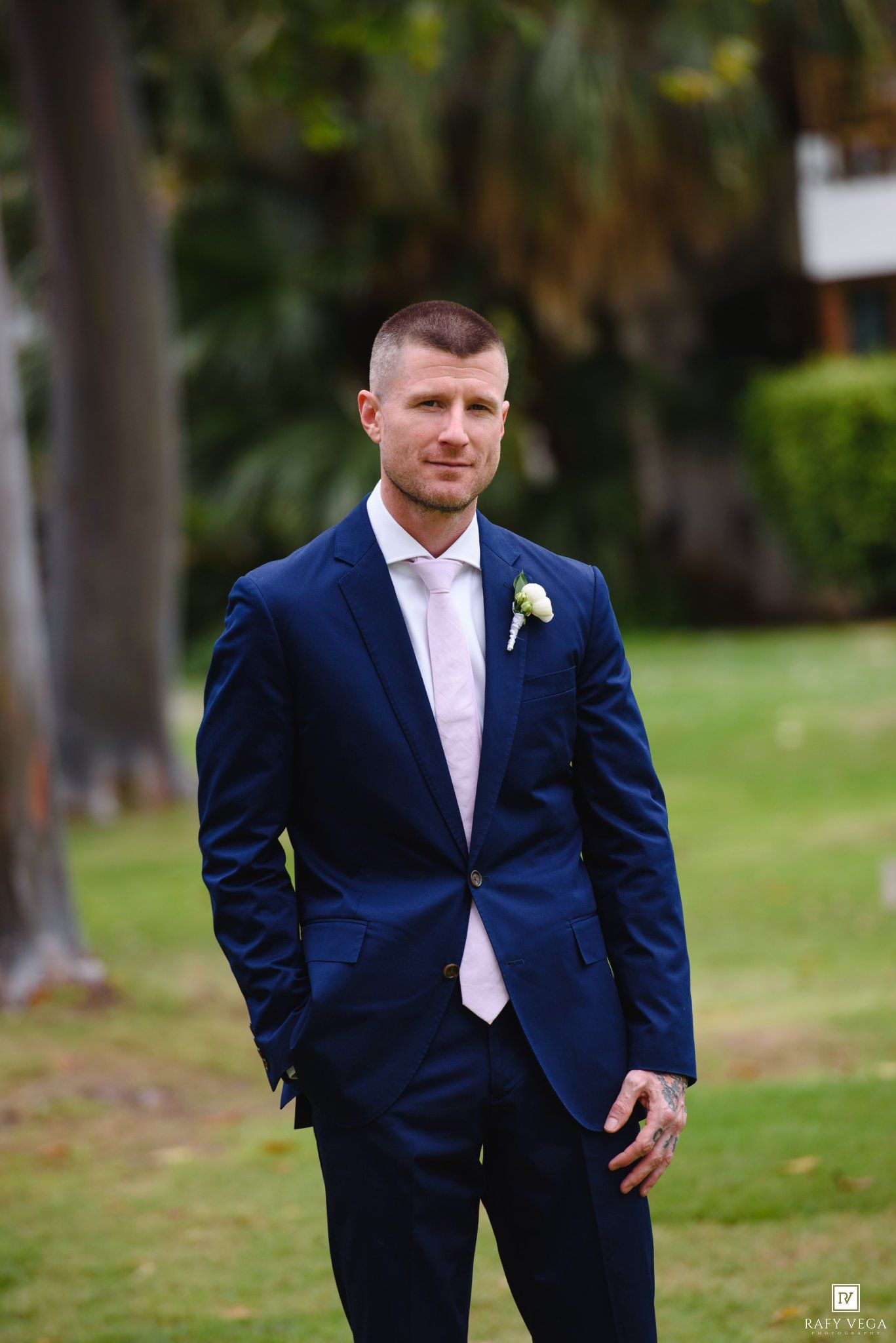 Groom in navy blue suit with pink tie and boutonniere posing outdoors on green lawn at wedding.