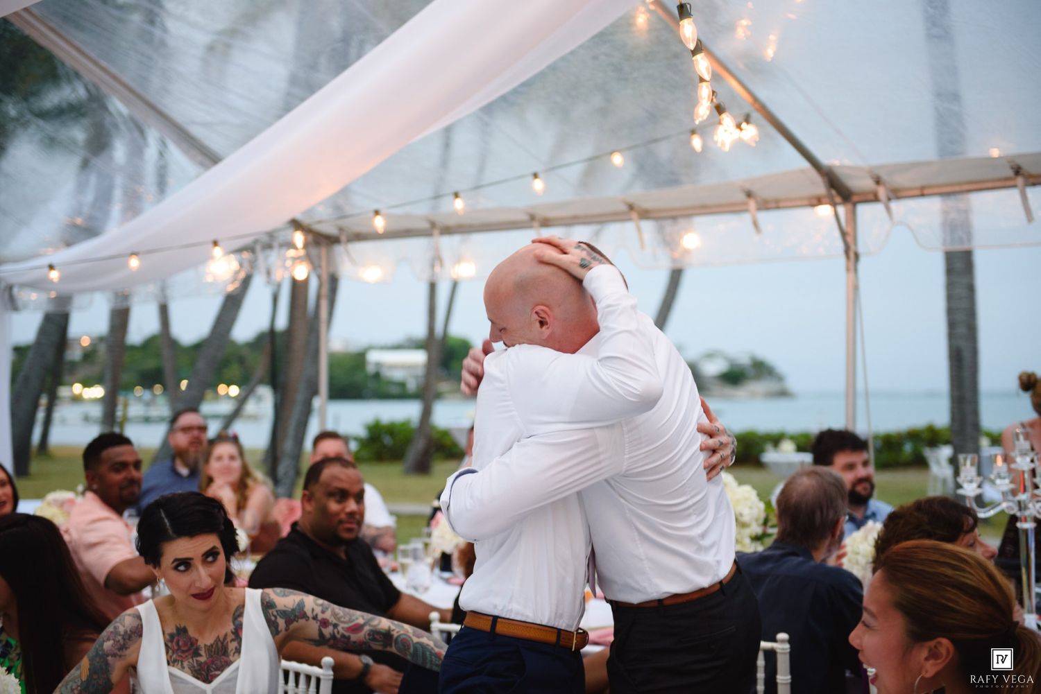 Two men share an emotional hug at an outdoor wedding reception under a white tent with string lights.