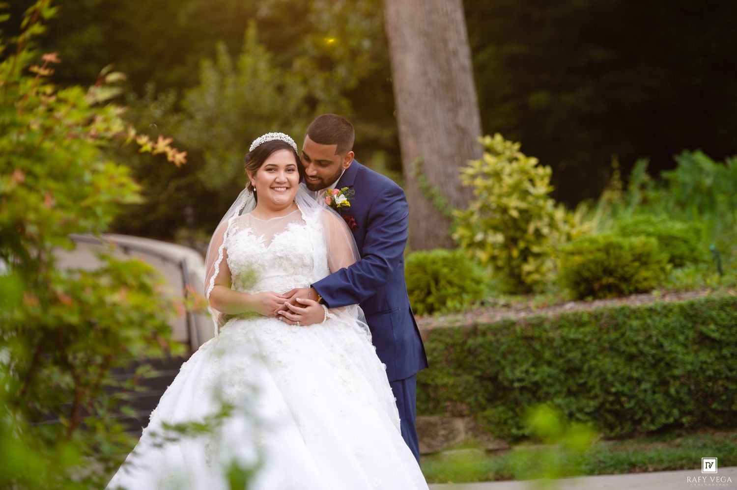 Bride and groom embracing in lush garden, bride in white ball gown and groom in navy suit.