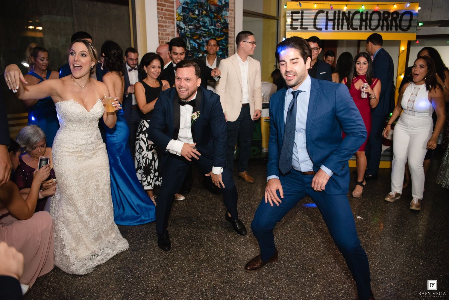 Wedding guests dance energetically at El Chinchorro venue, bride in white gown celebrating with groom and friends.
