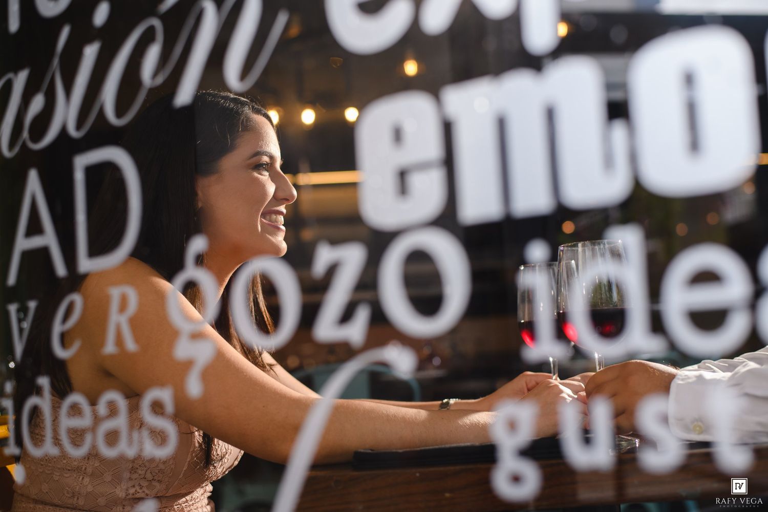 Woman smiling and holding a champagne glass at a bar with decorative text wall behind her.