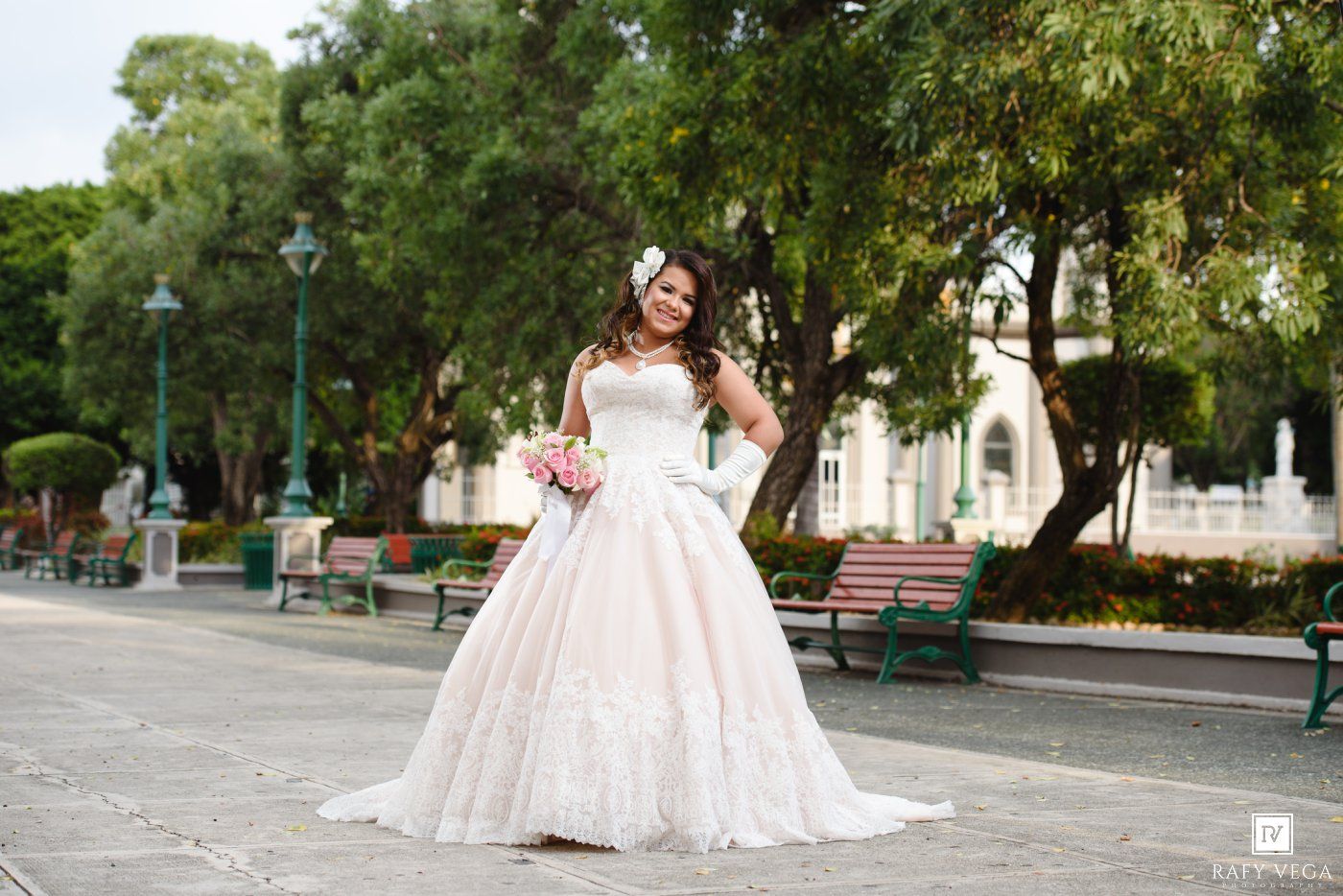 Bride in elegant white ball gown holding pink bouquet poses outdoors in park with green trees and benches.