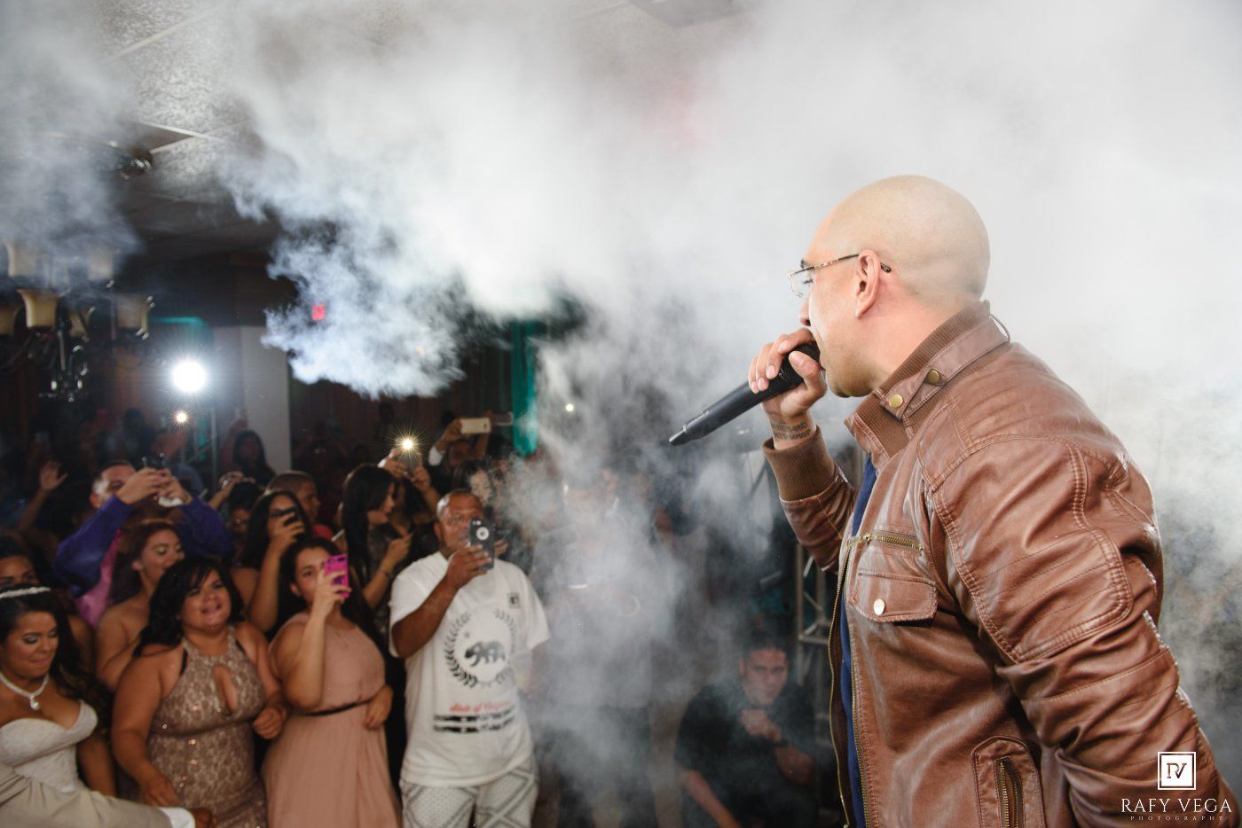 Bald male performer singing into microphone on stage with smoke effects and crowd at an outdoor night event.