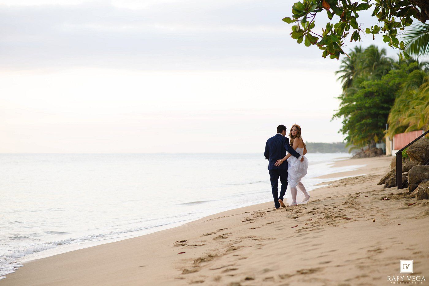 Couple walking hand in hand along a tropical beach at sunset with lush green trees and calm ocean waves.