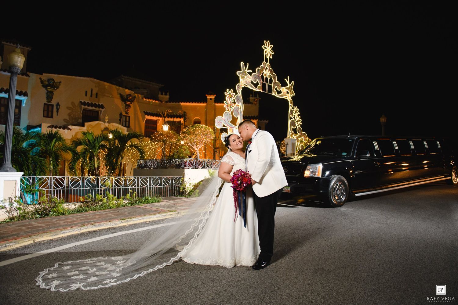 Bride and groom kissing outside at night near a stretch limo with festive lights glowing behind them.