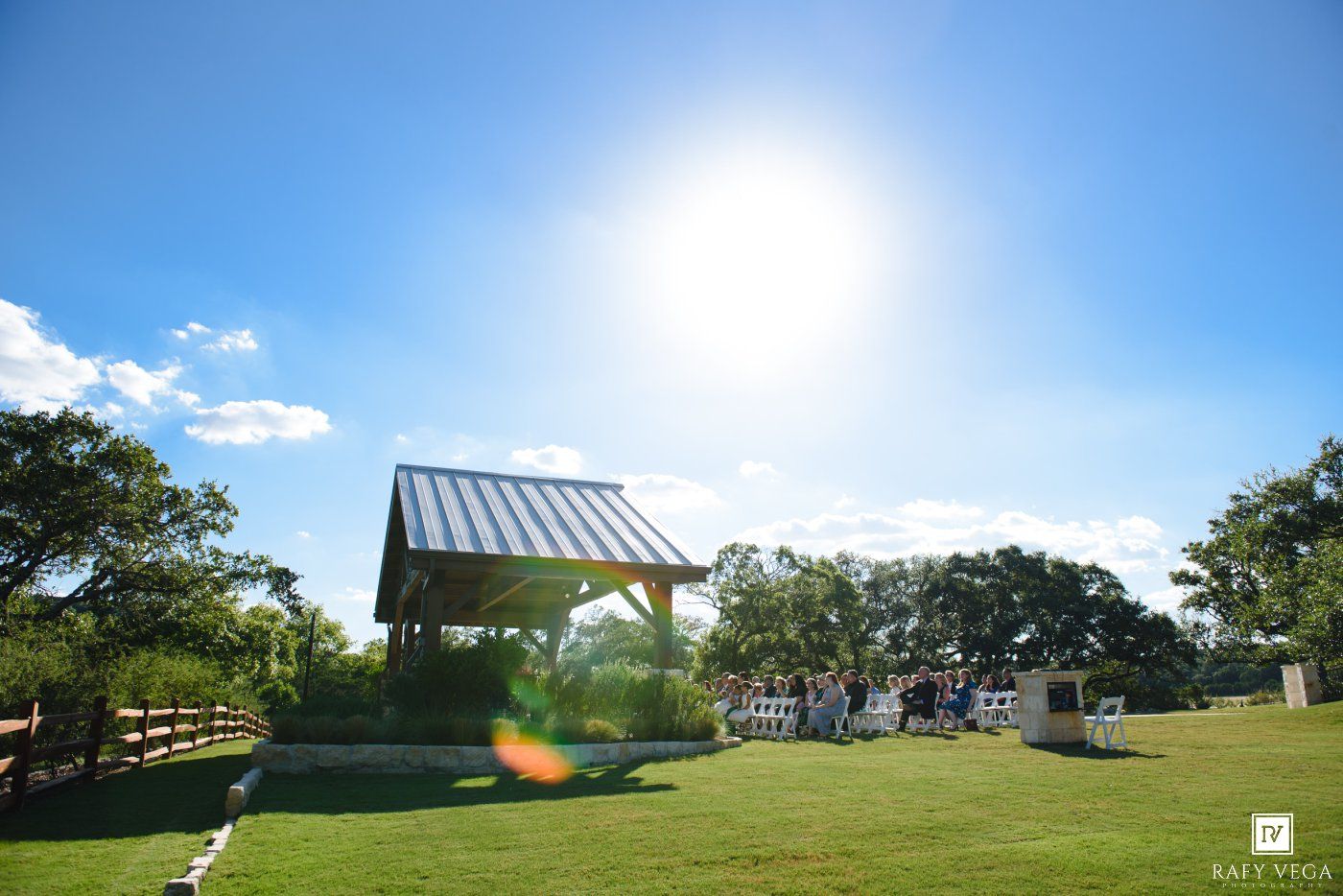 Sunny outdoor wedding venue with wooden pergola on lush green lawn surrounded by trees and white chairs under bright blue sky.