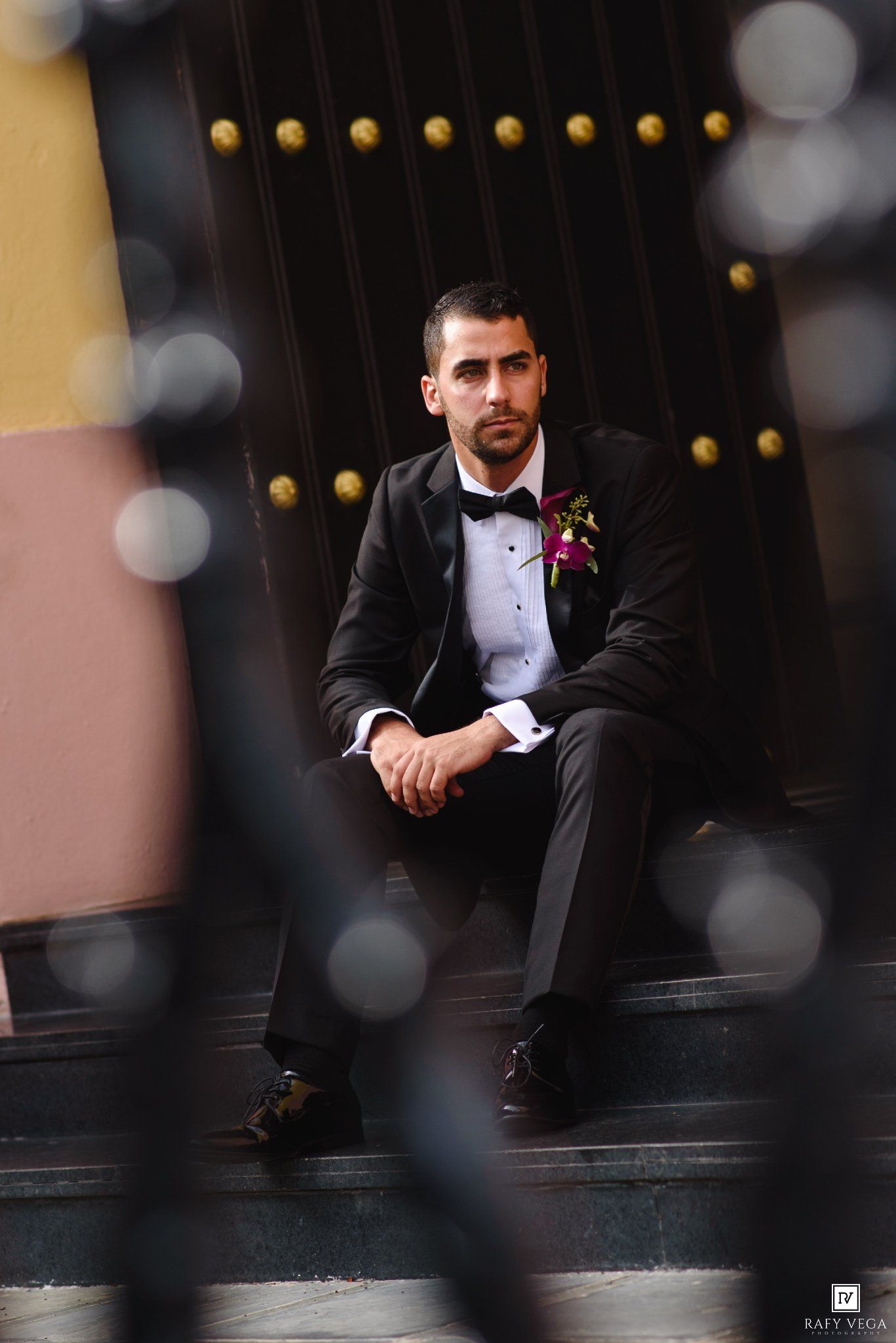 A groom in a black tuxedo with floral boutonniere sits on steps, photographed through a decorative iron fence.
