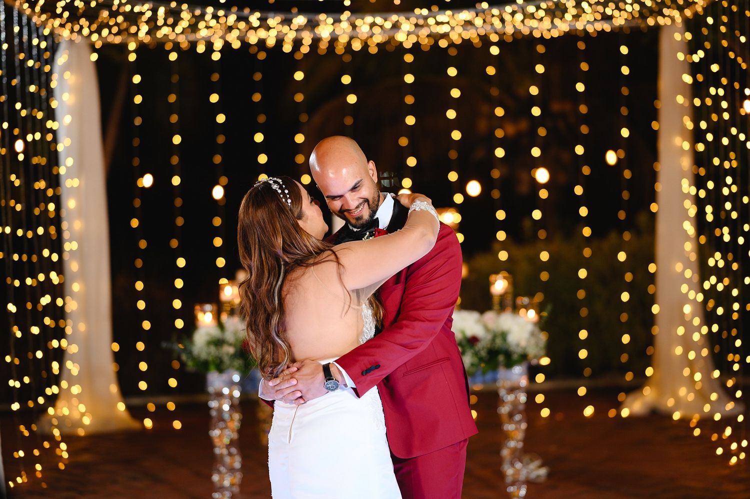 Newlyweds embracing during first dance beneath warm twinkling fairy light curtain at night.