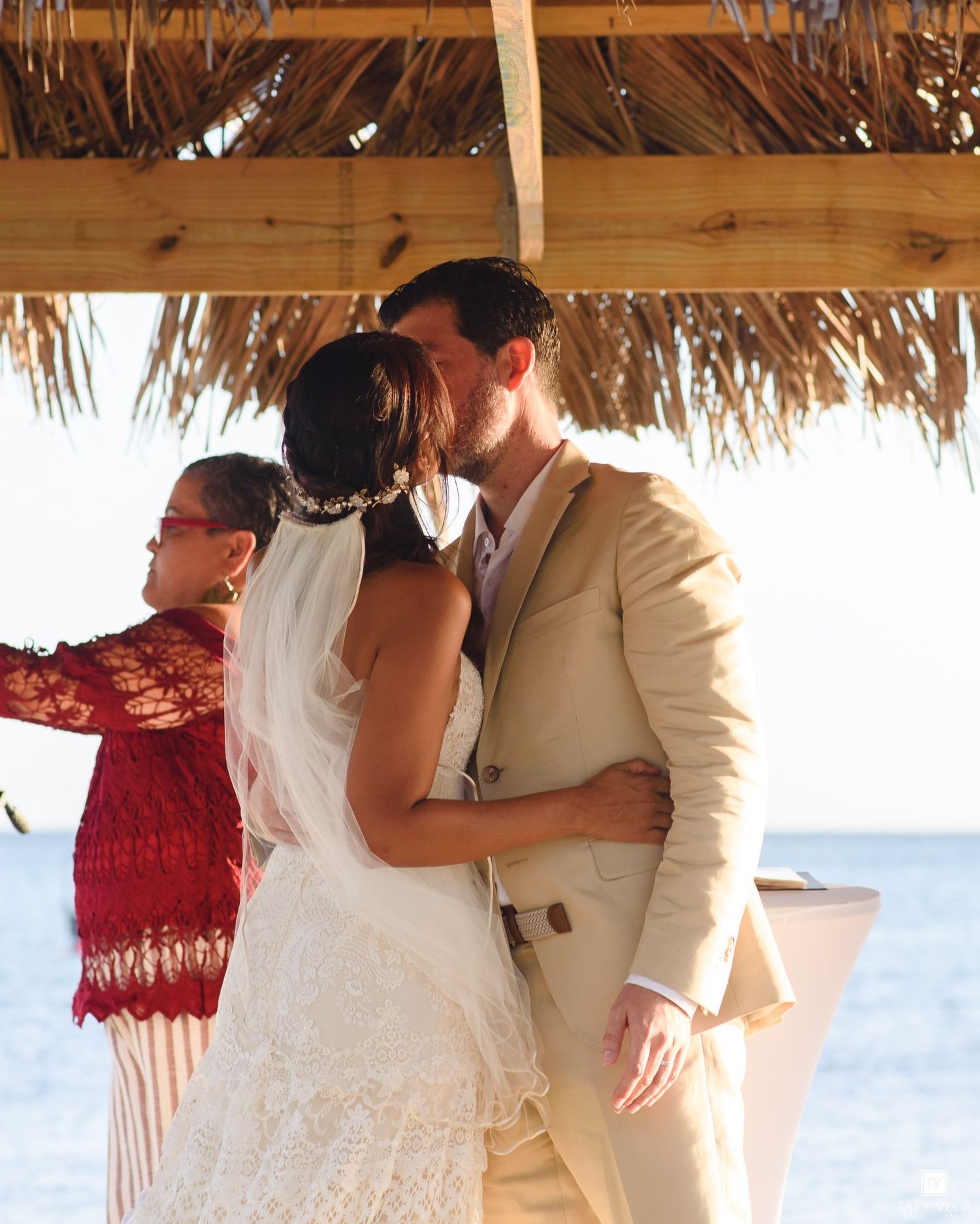 Bride and groom share first kiss at outdoor beach wedding ceremony under thatched roof with ocean backdrop.