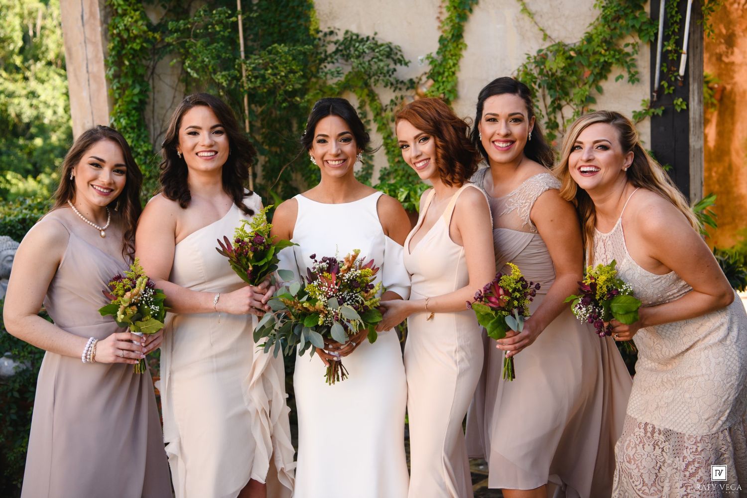 Bride in white gown poses with five bridesmaids in taupe dresses holding colorful bouquets outdoors.