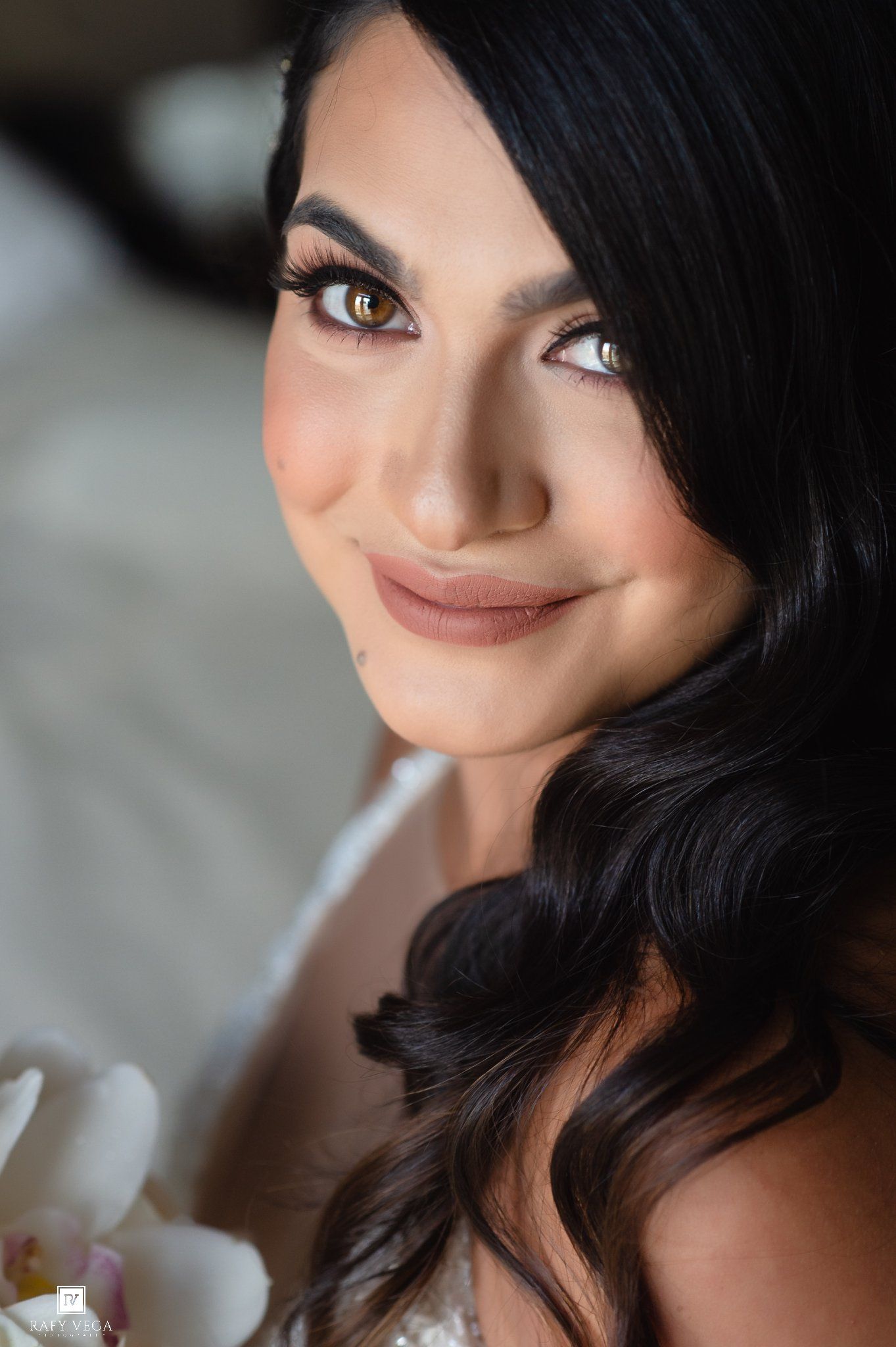 Close-up portrait of a bride with dark hair, golden eyes, and elegant makeup holding white flowers.
