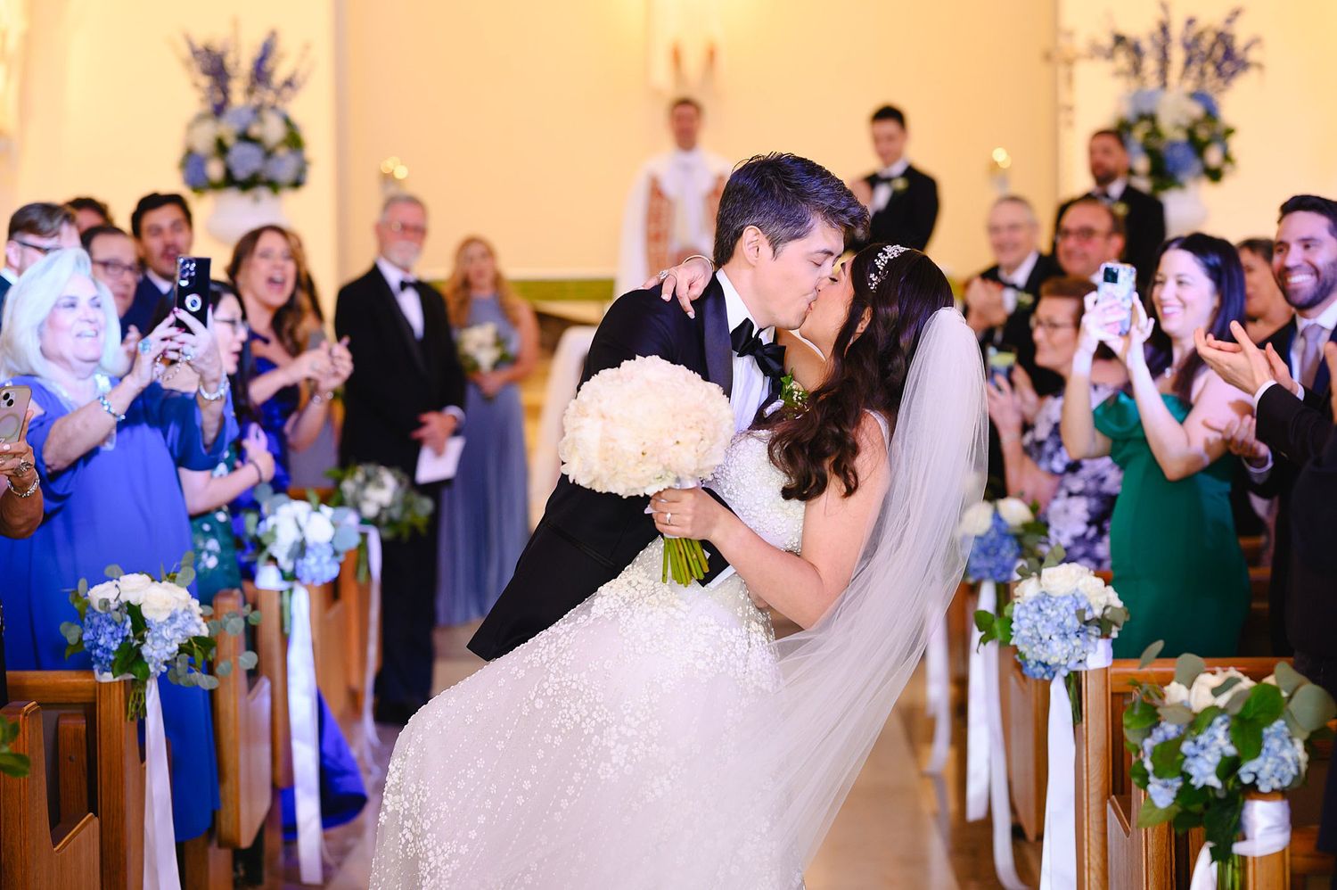Bride and groom share first kiss walking down church aisle surrounded by cheering guests at elegant wedding ceremony.