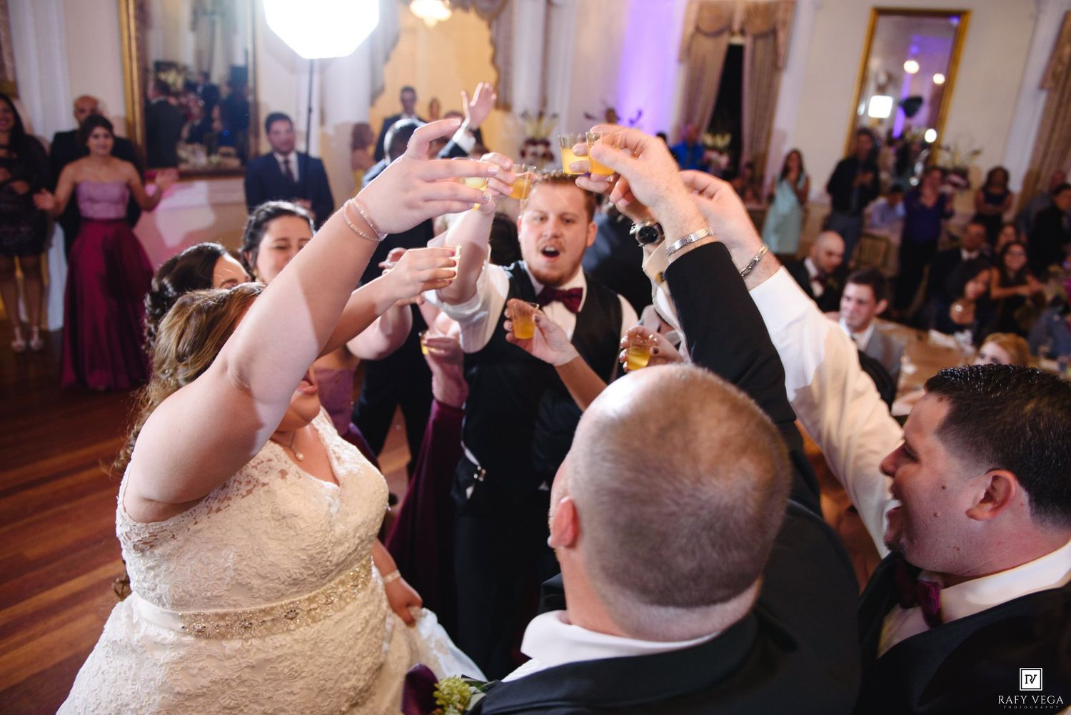 Wedding guests raise glasses in a joyful toast at an elegant reception hall, with bride and groom at center.