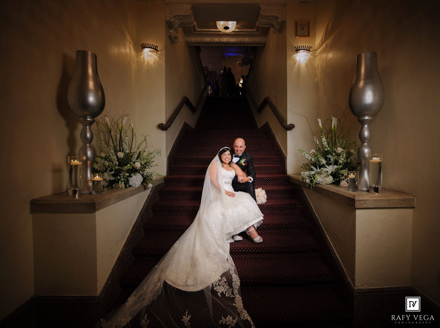 Bride and groom descend elegant staircase in warmly lit wedding venue, surrounded by floral arrangements.