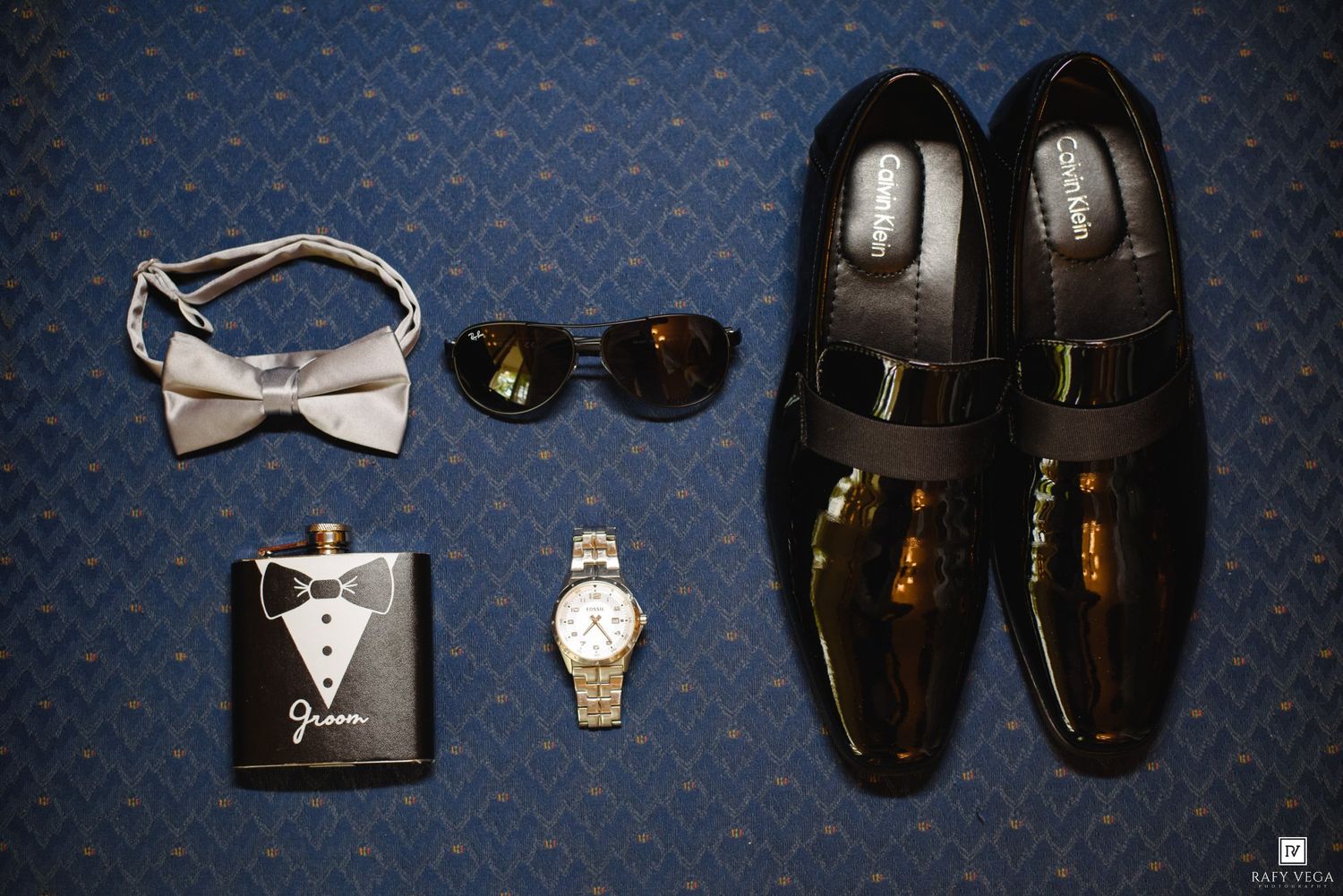 Groom accessories flatlay featuring patent leather shoes, bow tie, sunglasses, gold watch, and a tuxedo flask.