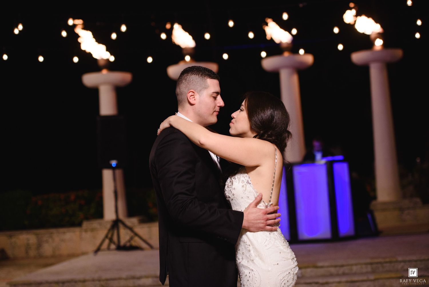 Couple shares romantic first dance outdoors at night wedding reception with torch lights and string lights in background.