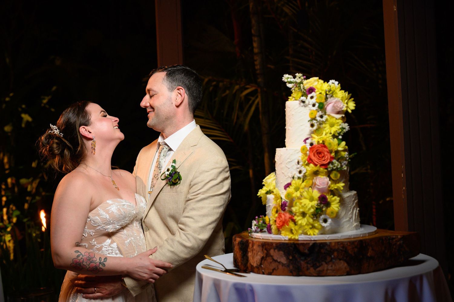 Bride and groom smiling together beside a colorful floral wedding cake at an outdoor evening reception.