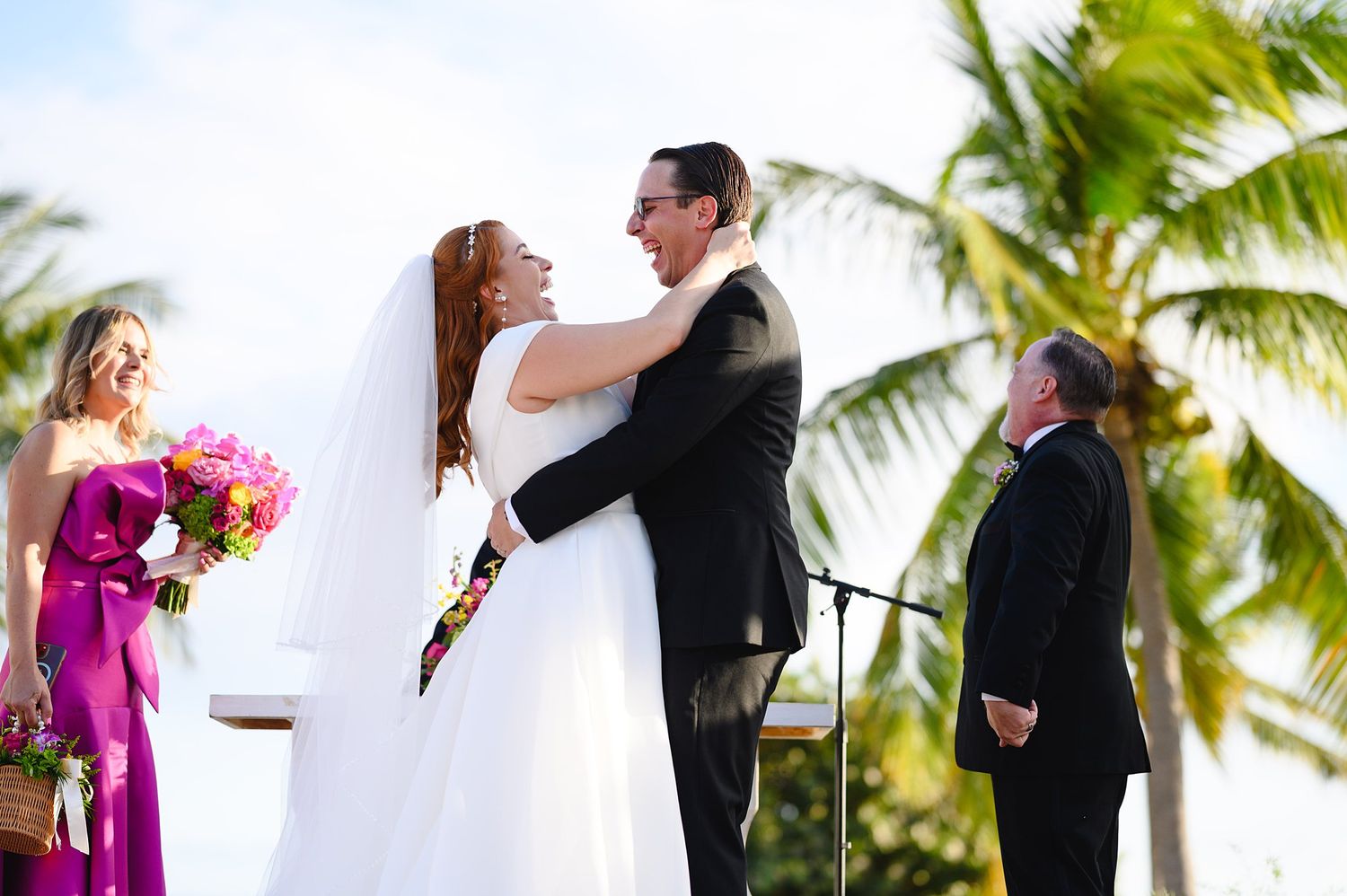 Bride and groom share first embrace after wedding vows at tropical outdoor ceremony with palm trees and pink flowers.