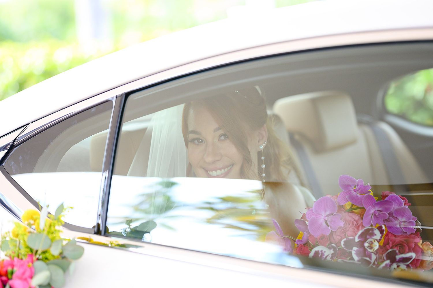 Smiling bride in wedding veil sitting in decorated car with pink orchids and yellow flowers on wedding day.