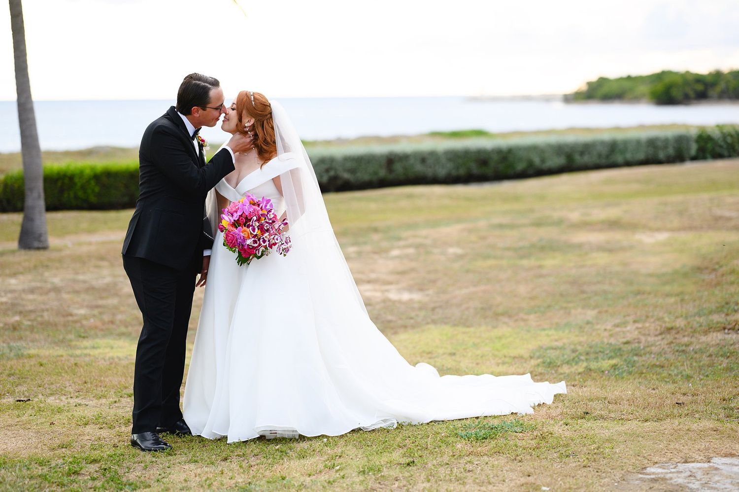 Bride and groom sharing a kiss outdoors near the ocean, bride holding a vibrant pink bouquet in a flowing white gown.