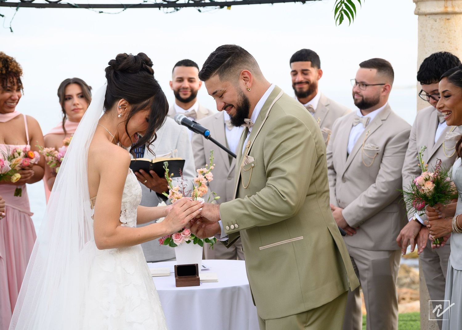 Bride in a white gown and groom in a tan suit exchange rings at an outdoor wedding ceremony as smiling groomsmen and bridesmaids look on.