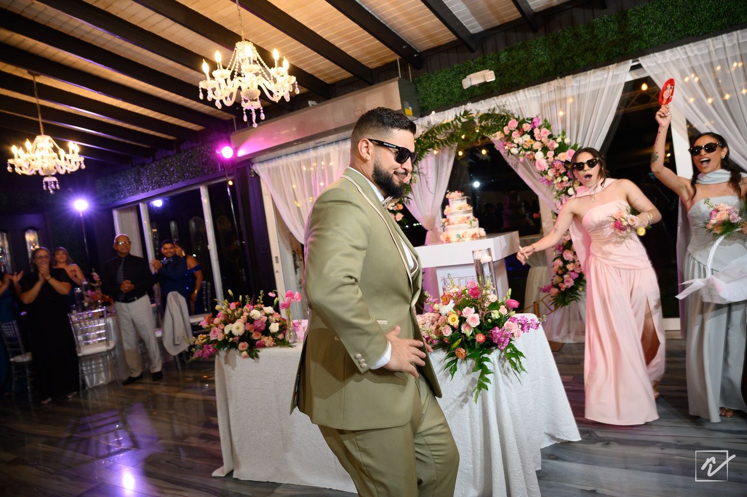 Groom in an olive green suit and sunglasses dances at a wedding reception as bridesmaids in pink dresses cheer beside floral tables.