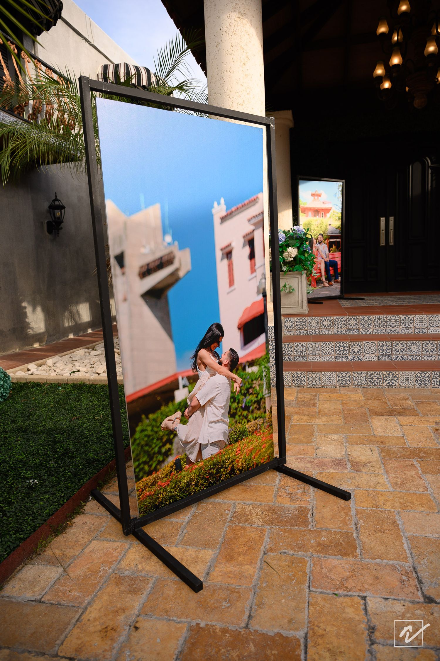 Large framed pre-wedding photo of a couple embracing in a garden, displayed on an easel on a terracotta tiled patio at a venue entrance.