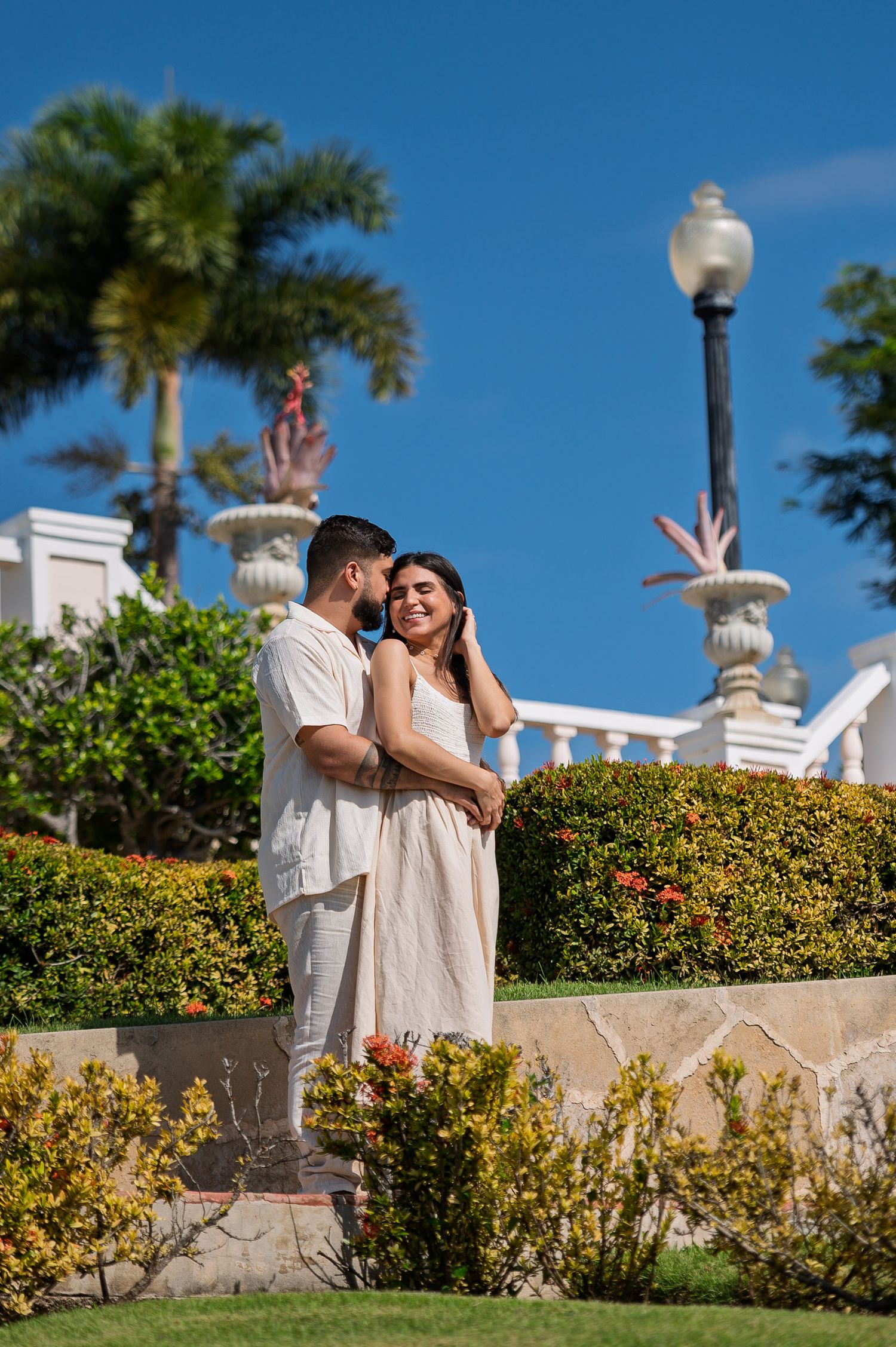 A couple in matching beige outfits embracing in a lush garden with palm trees and blue sky.