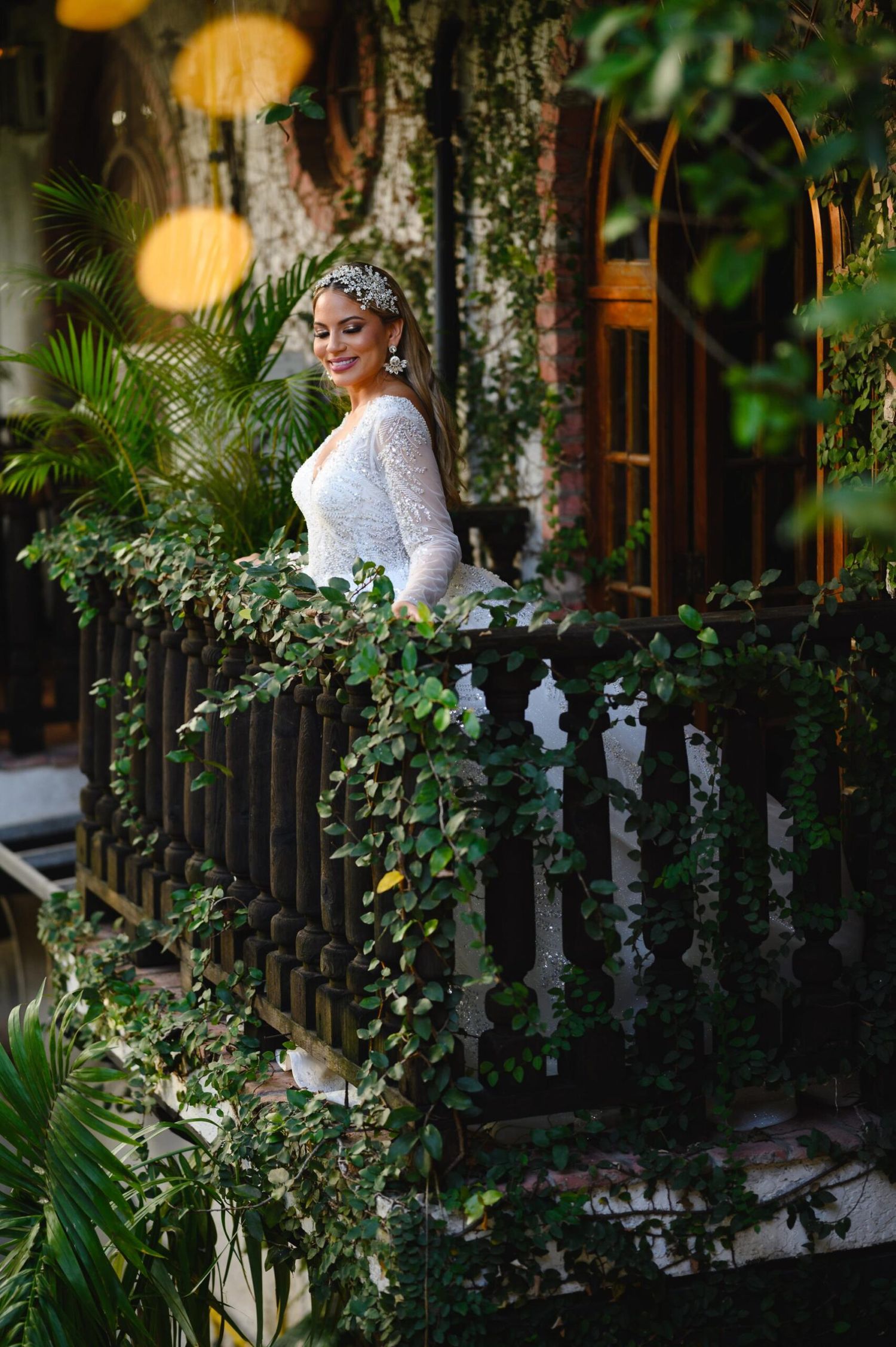 Bride in white lace gown and tiara poses on ivy-covered balcony surrounded by lush tropical greenery.