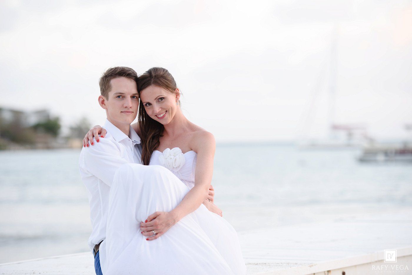 A happy couple in white wedding attire embraces on a beach pier with a sailboat and ocean in the background.