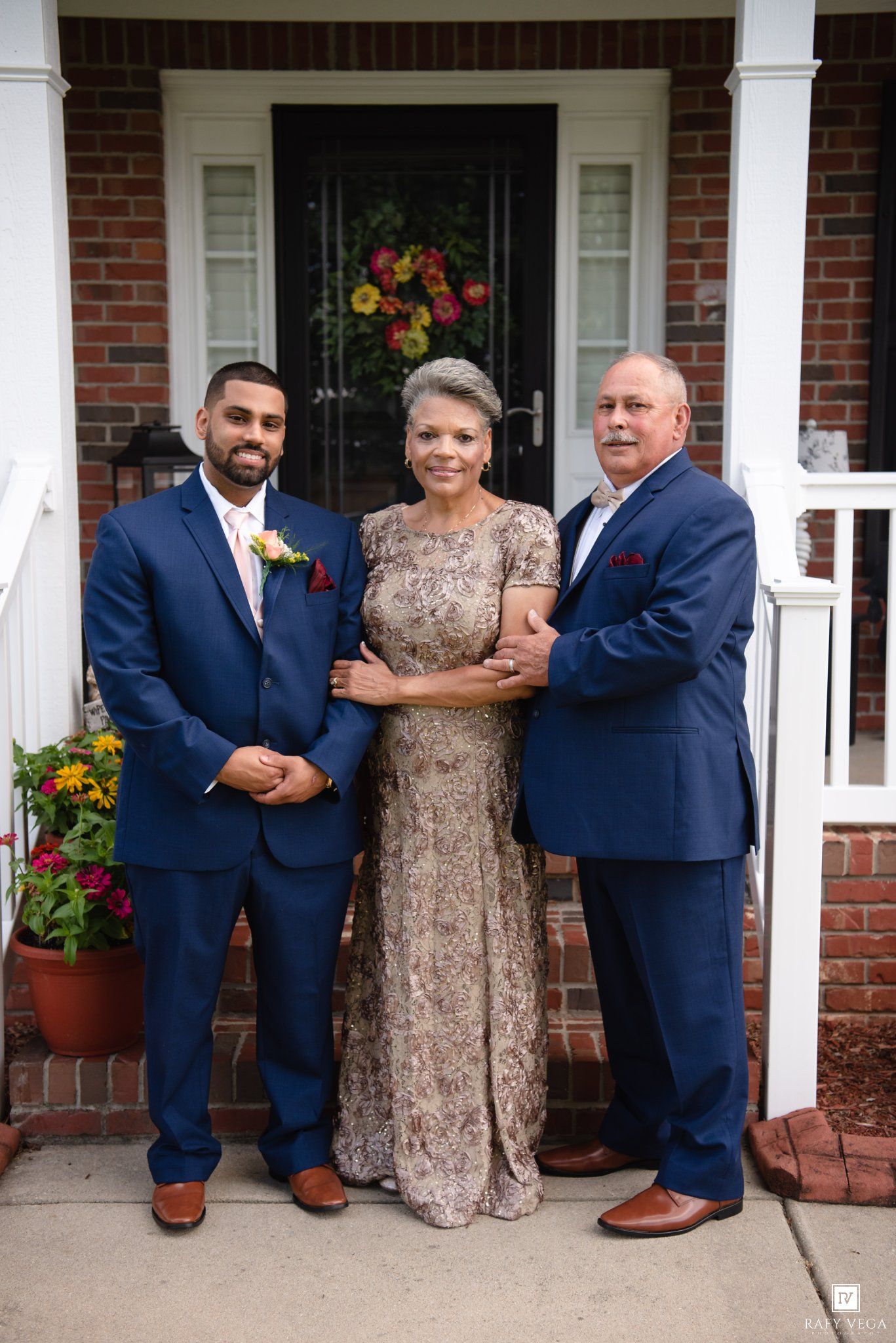 Two men in navy suits and a woman in a gold gown pose together on a brick home's front steps.