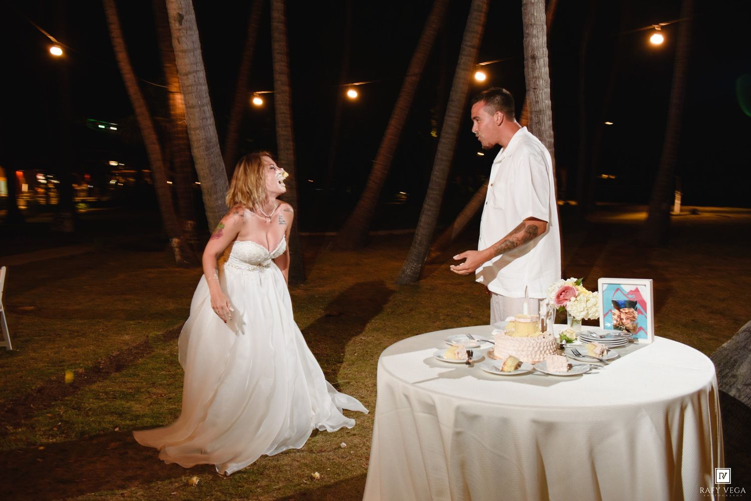 Bride and groom cutting wedding cake outdoors at night under palm trees with string lights at tropical venue.
