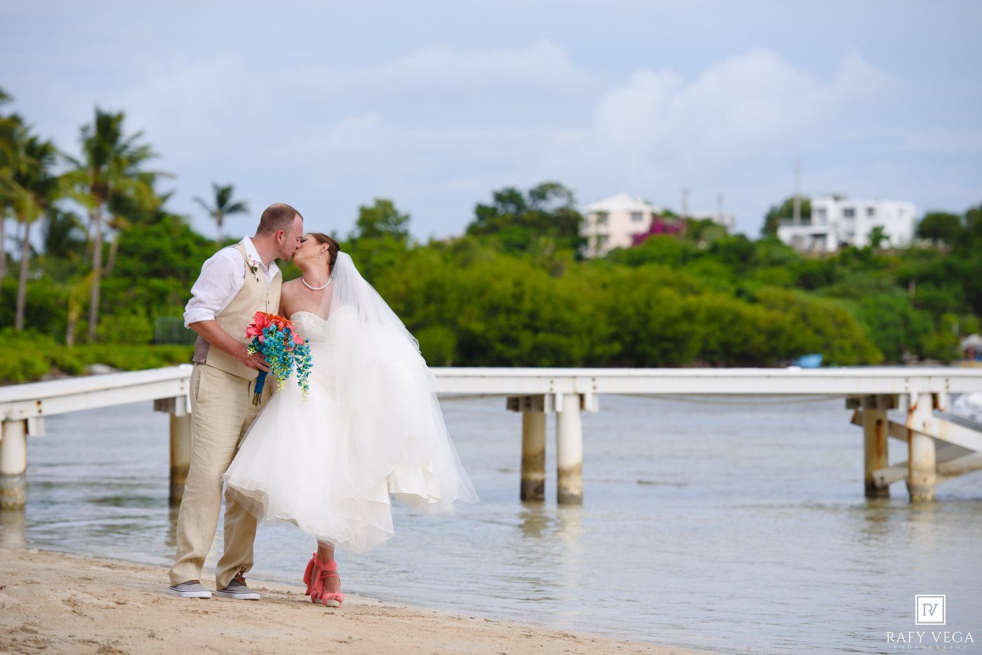 Bride and groom sharing a romantic kiss on a sandy beach near a wooden pier during their tropical wedding.