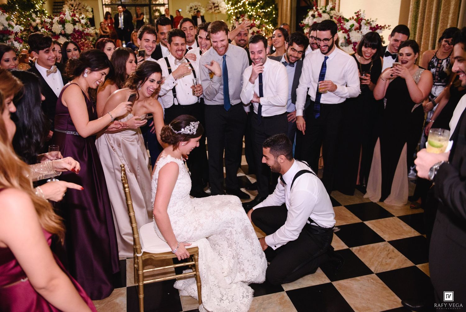 Groom removes garter from bride seated on chair at wedding reception while guests cheer on black and white checkered dance floor.