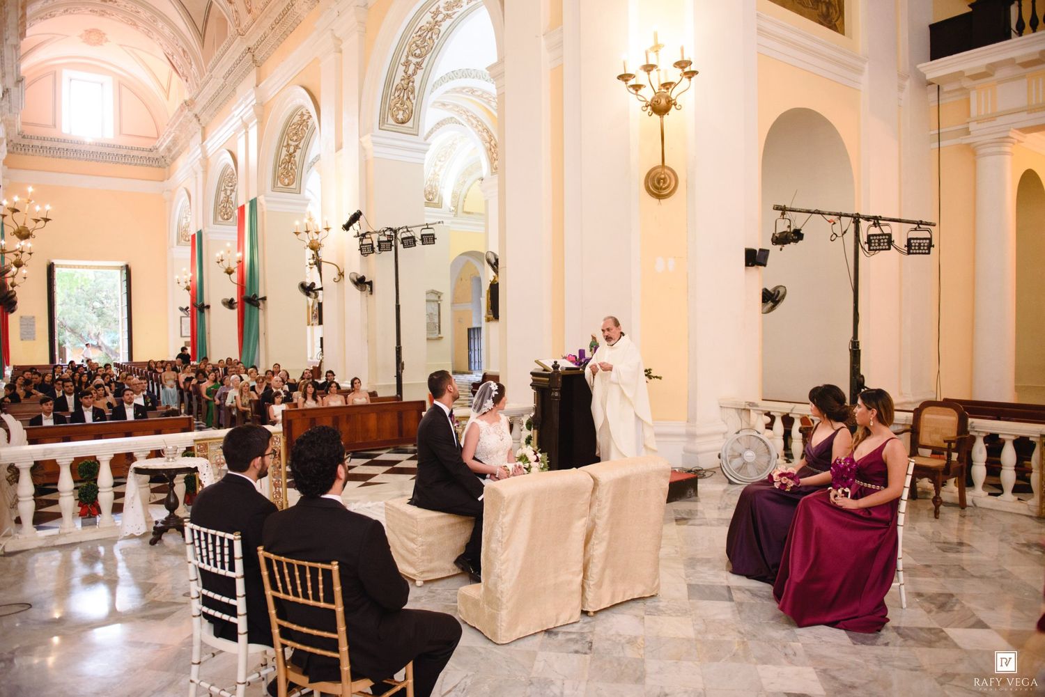 Elegant wedding ceremony inside a grand church with ornate columns, guests seated, and bridal party gathered at the altar.